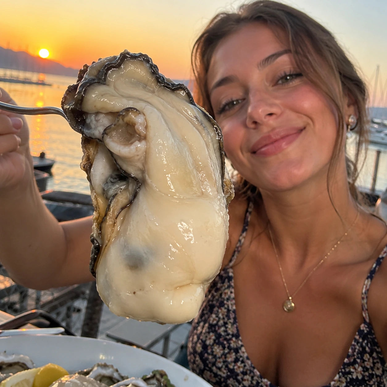 Woman enjoying oysters at a waterfront table at sunset.